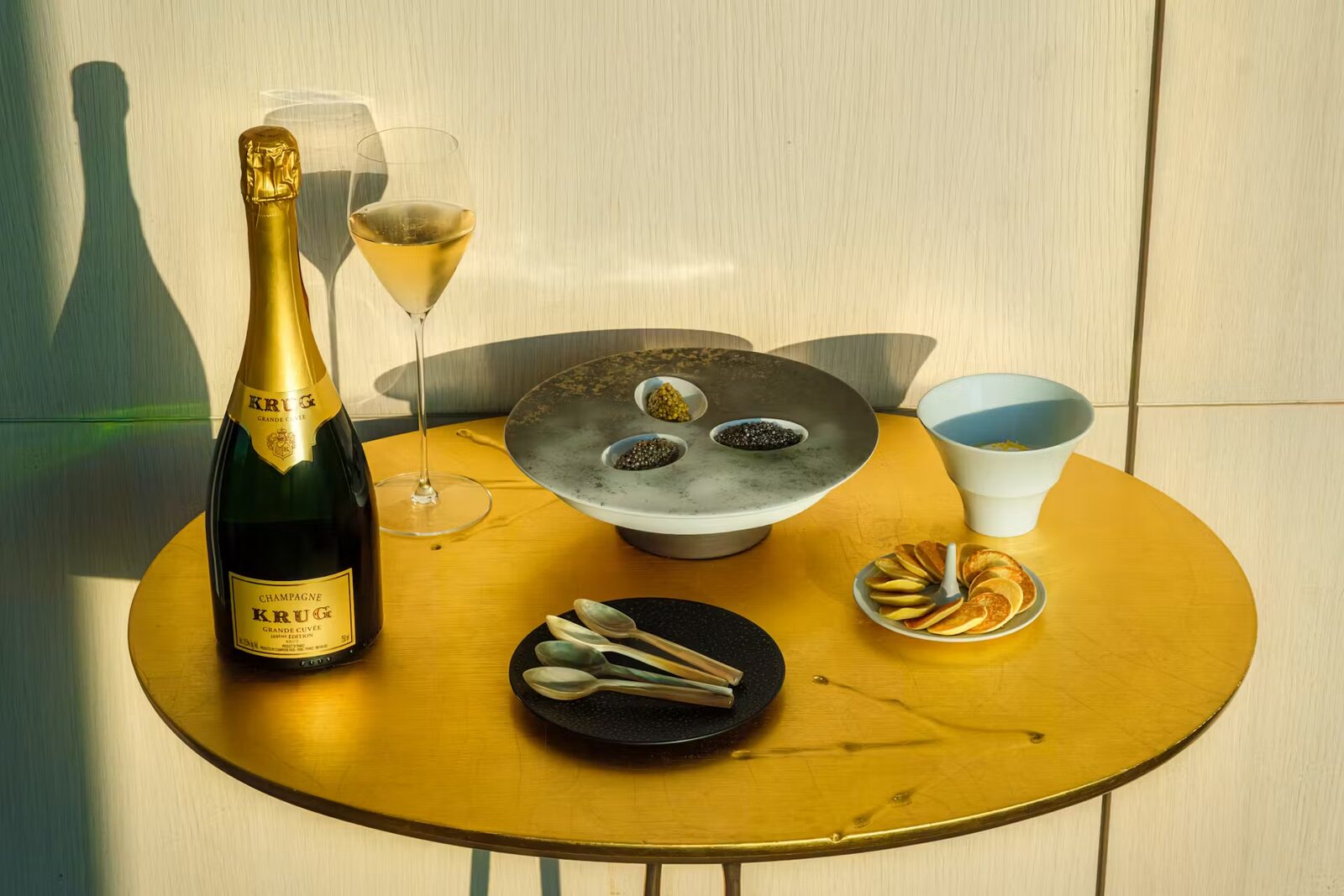 A gilded café table set with Krug champagne, a poured glass, three caviar tins on a stone dish, mother-of-pearl spoons and blini