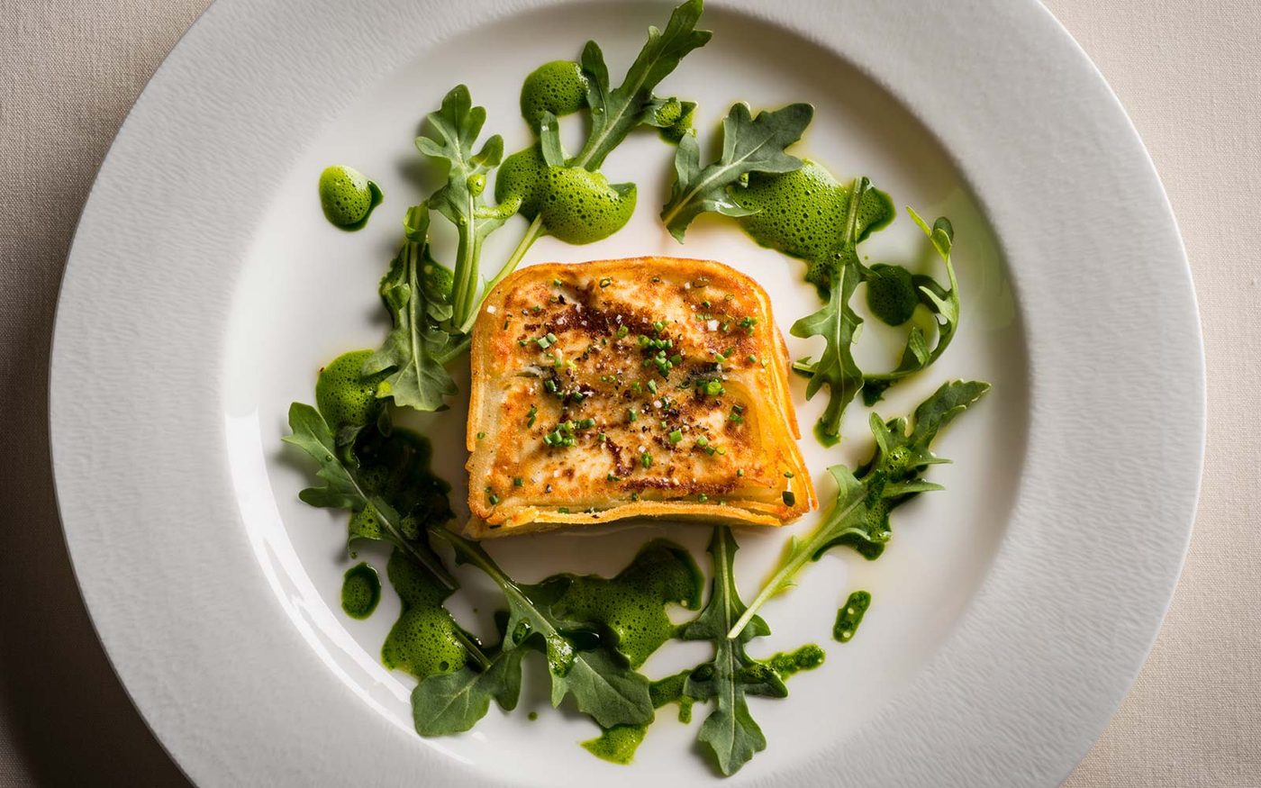 A golden-crusted potato and cheese gratin square with chives, arugula and a herb oil foam on a white plate