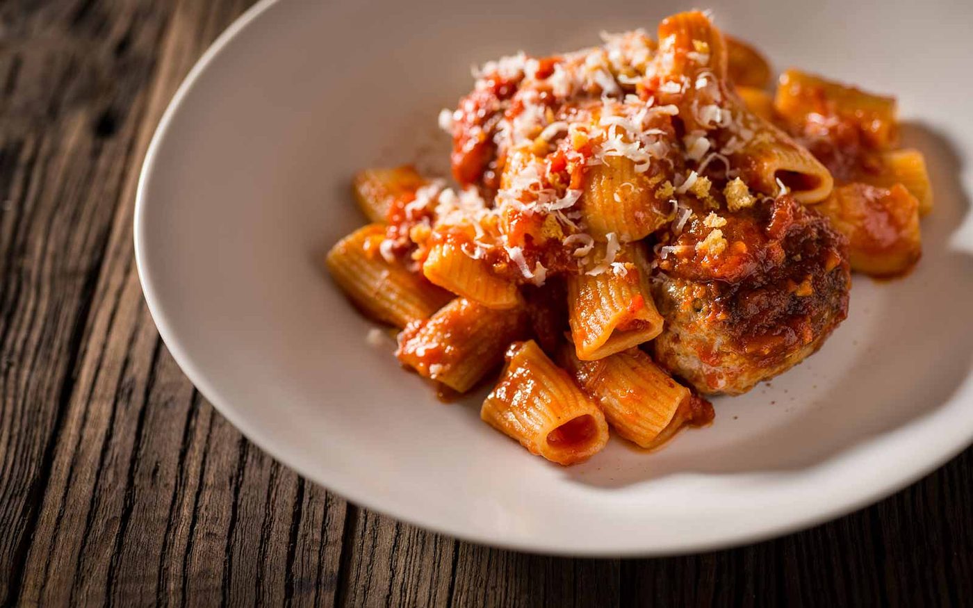 Rigatoni in tomato sugo with a house meatball, grated pecorino and toasted breadcrumbs, in a wide white bowl on reclaimed wood