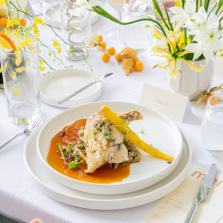 A plated chicken course with pan jus and micro greens on a set table with white linen, spring flowers and a handwritten place card