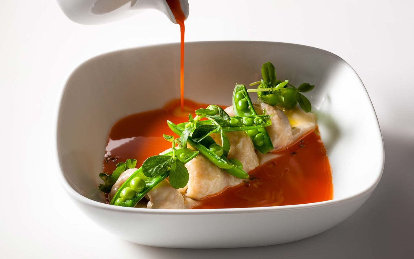 Poached white fish quenelles with sweet peas and pea shoots in a shallow bowl, with a red pepper broth being poured over at the table