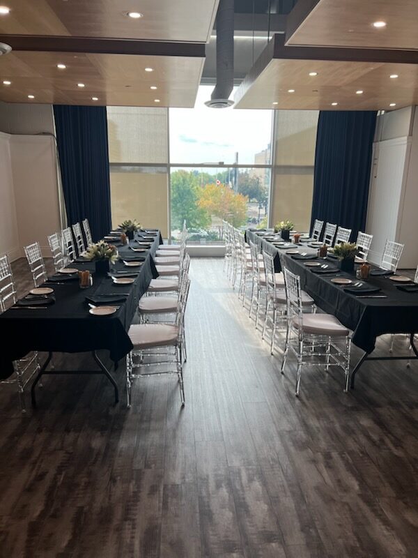 Two long banquet tables facing a window wall, set with black linen, greenery and candles
