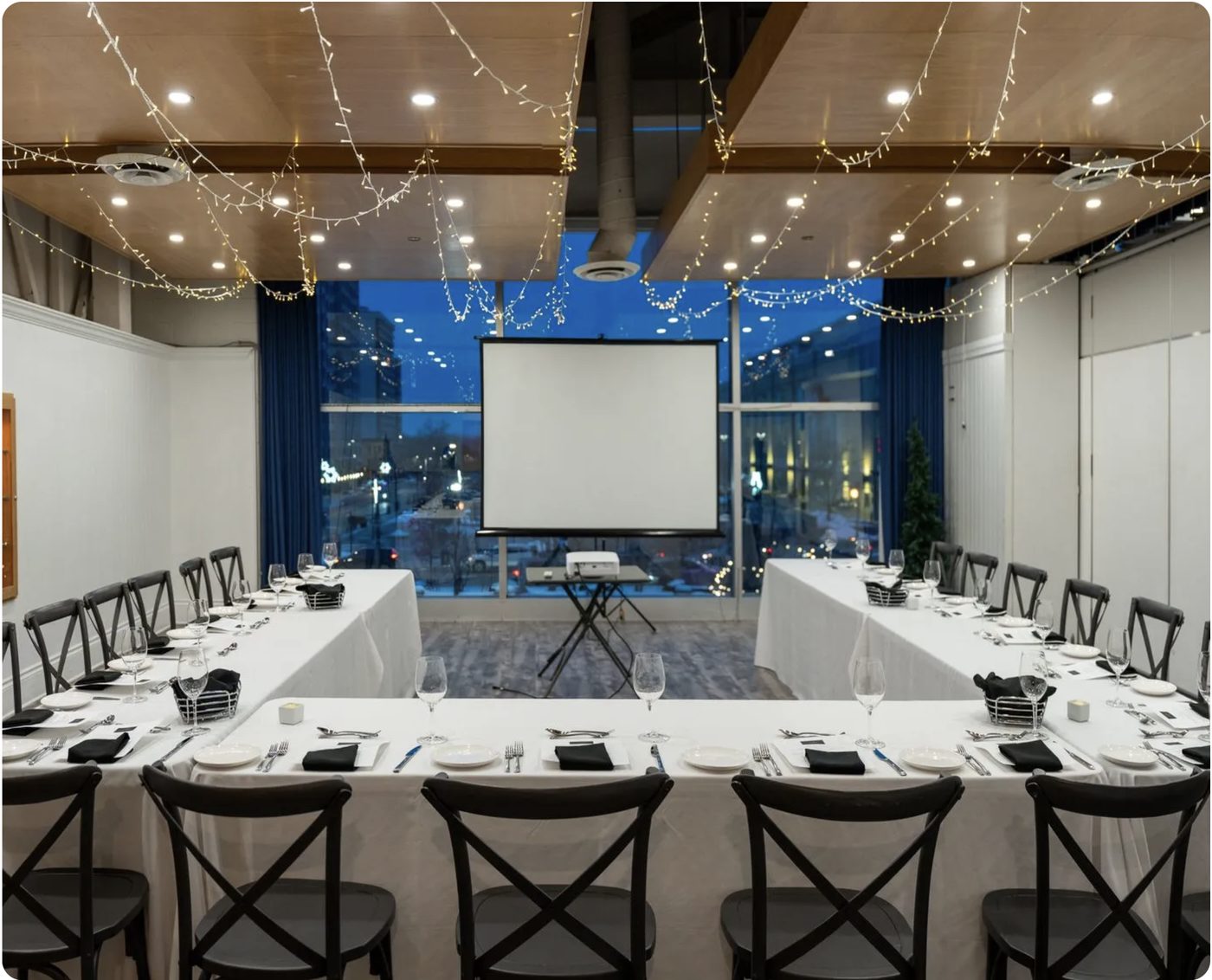 A U-shaped banquet table set with white linen and black napkins under strings of fairy lights, facing a projector screen and a window wall over downtown London at dusk