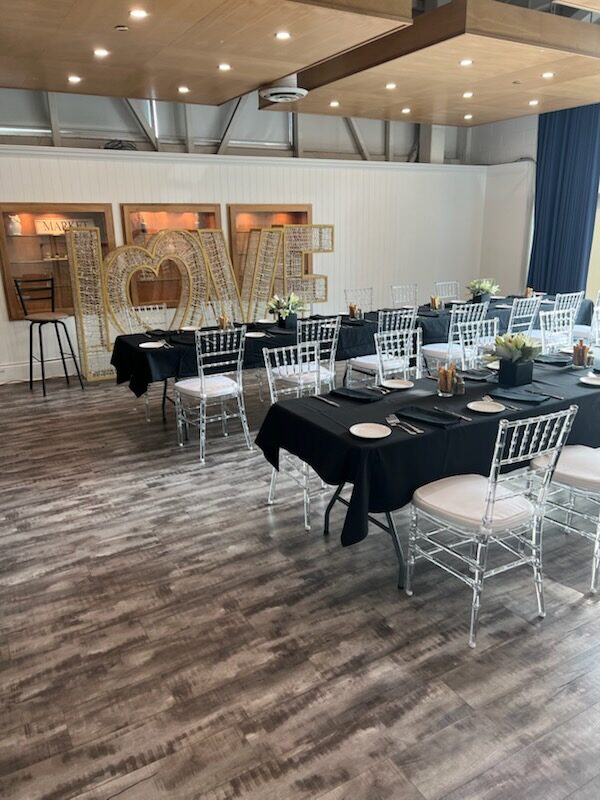 Event tables set with black linen and clear chiavari chairs beside an illuminated LOVE marquee sign