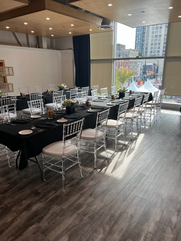 A long banquet table dressed in black linen with clear chiavari chairs, beside a floor-to-ceiling window looking over downtown London
